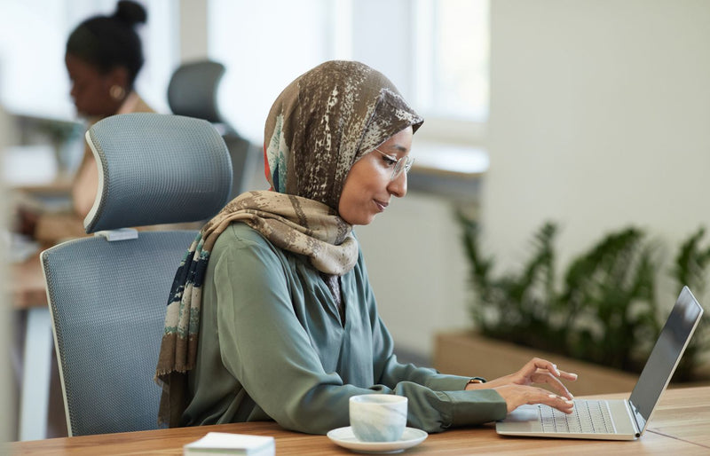 Middle-aged woman in hijab using laptop in modern office setting.