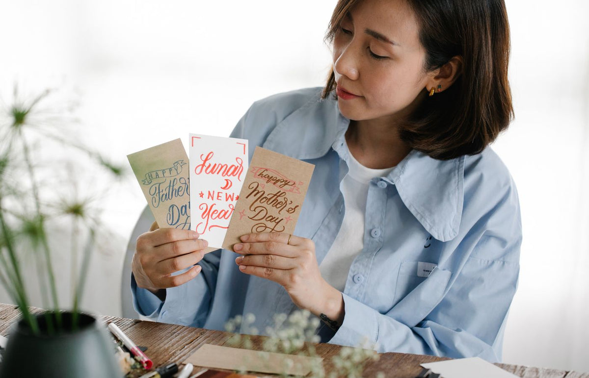 A woman in a blue shirt holds handmade greeting cards for Father's Day, Mother's Day, and Lunar New Year.
