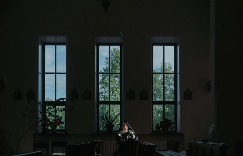 A nun reads quietly in a dimly lit church, illuminated by soft window light, epitomizing peaceful spirituality.