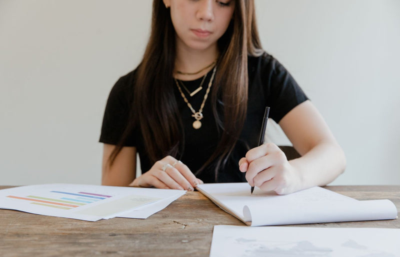 A woman focuses on writing while sitting at a wooden table, surrounded by papers. Bright indoor setting.