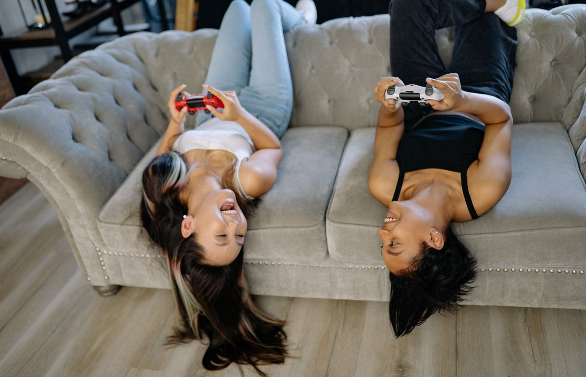 Two women having fun playing video games upside down on a sofa indoors.