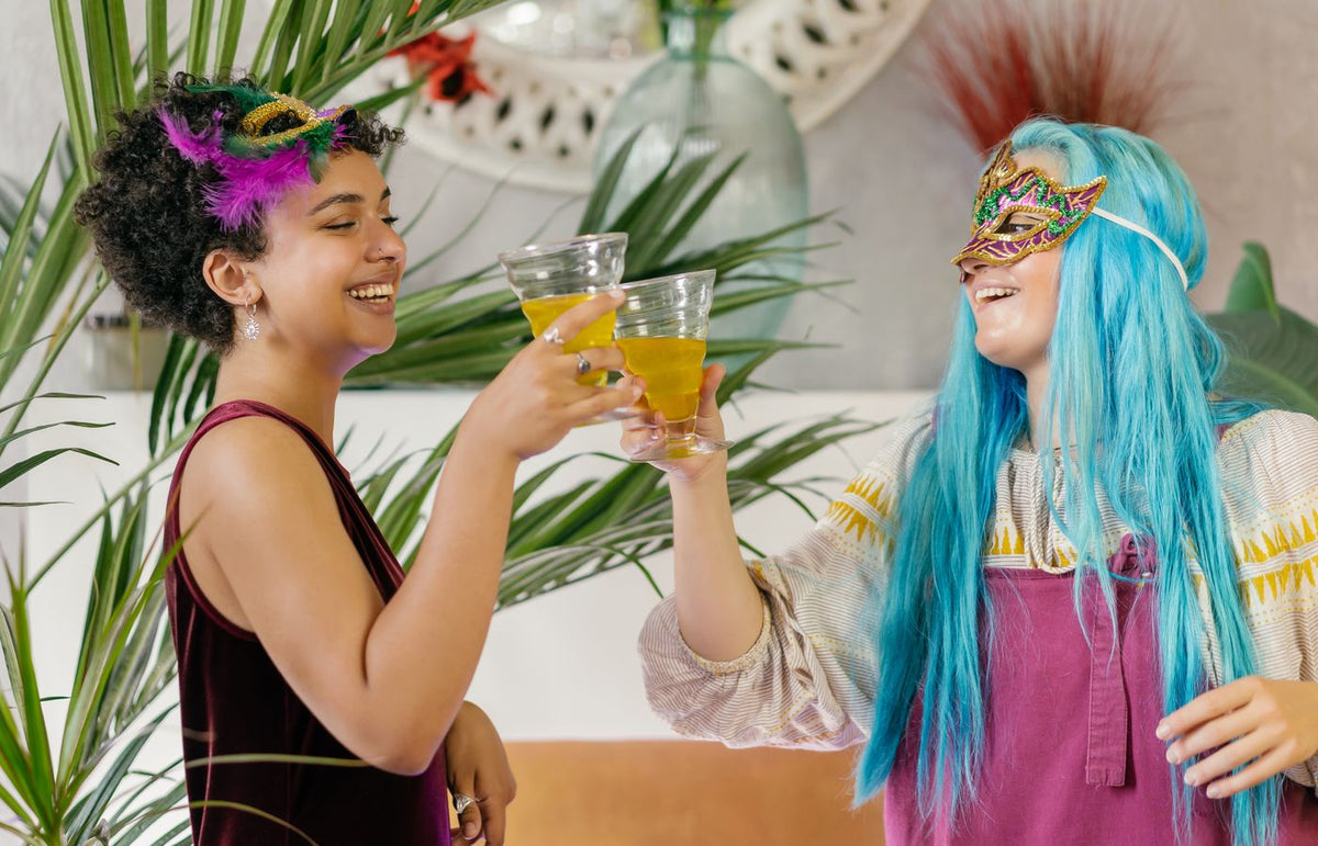 Two women enjoying a masquerade party, sharing drinks and laughter indoors.