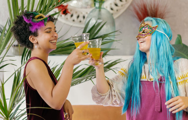 Two women enjoying a masquerade party, sharing drinks and laughter indoors.