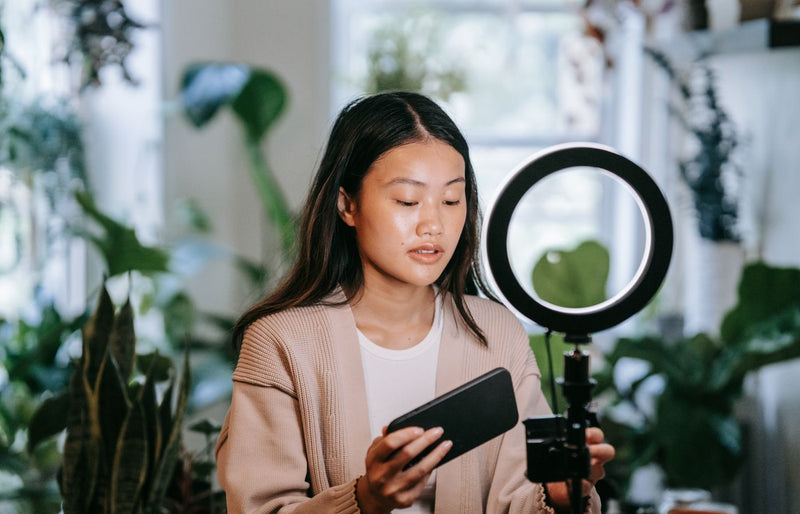 Asian woman adjusting smartphone and ring light indoors for video recording.