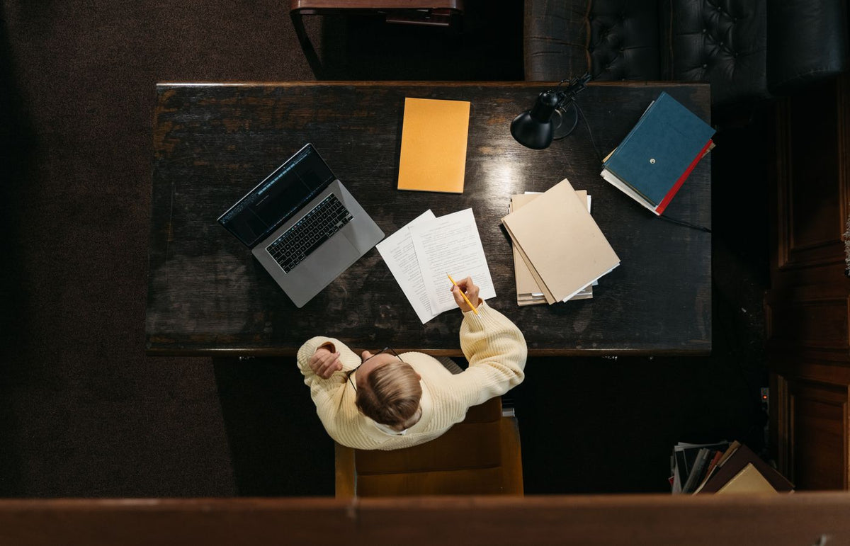 Person in yellow sweater working at a wooden desk with documents, folders, and a laptop.