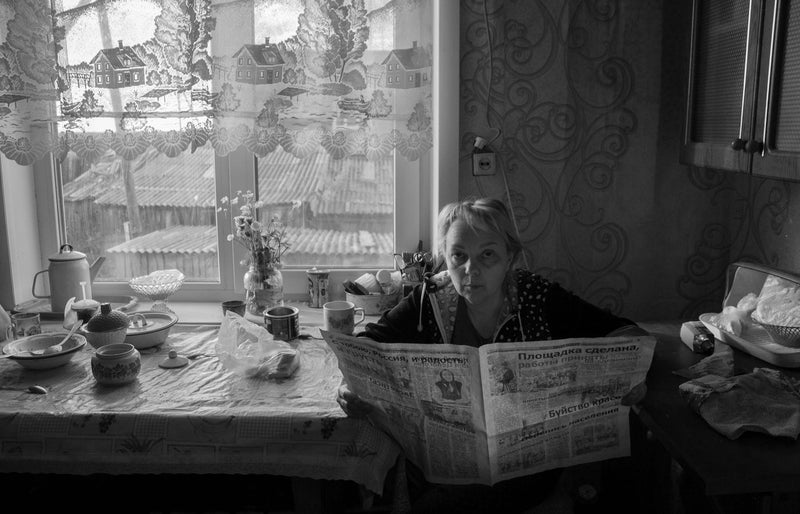 Black and white photo of an elderly woman reading a newspaper in a cozy kitchen setting.
