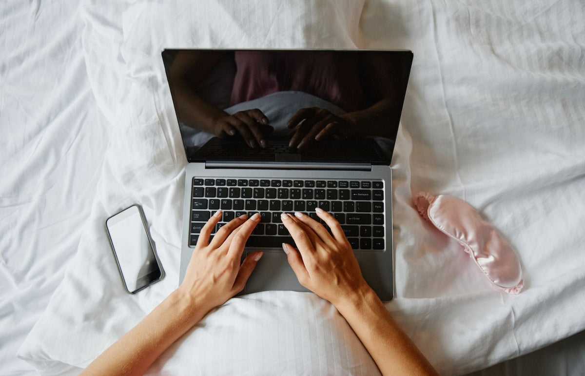 Person lying in bed using a laptop, with a phone and sleep mask nearby, creating a cozy indoor work environment.