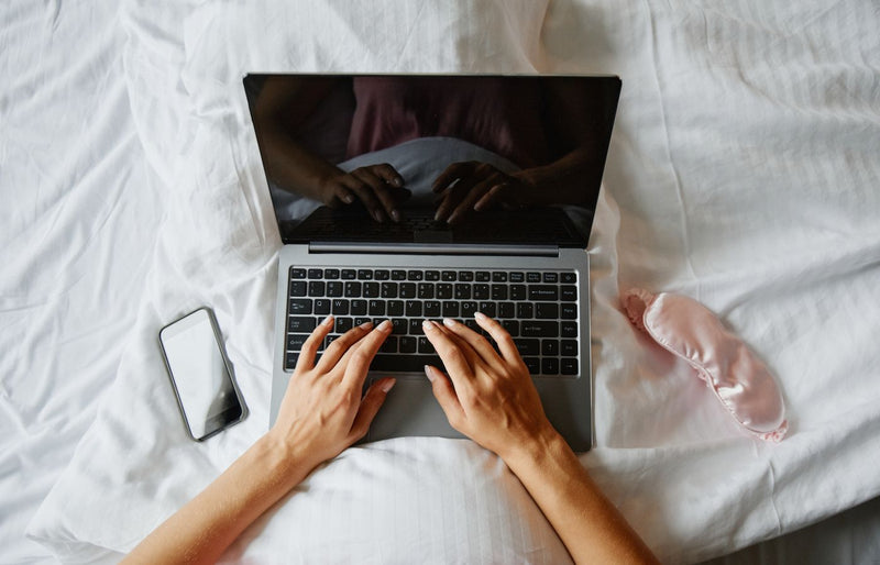 Person lying in bed using a laptop, with a phone and sleep mask nearby, creating a cozy indoor work environment.