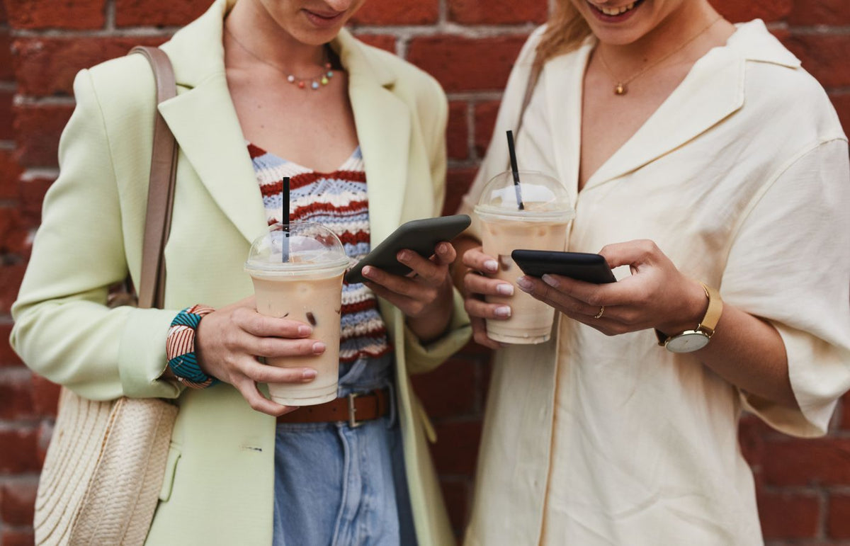 Two women enjoying iced coffee while browsing smartphones against a red brick wall.