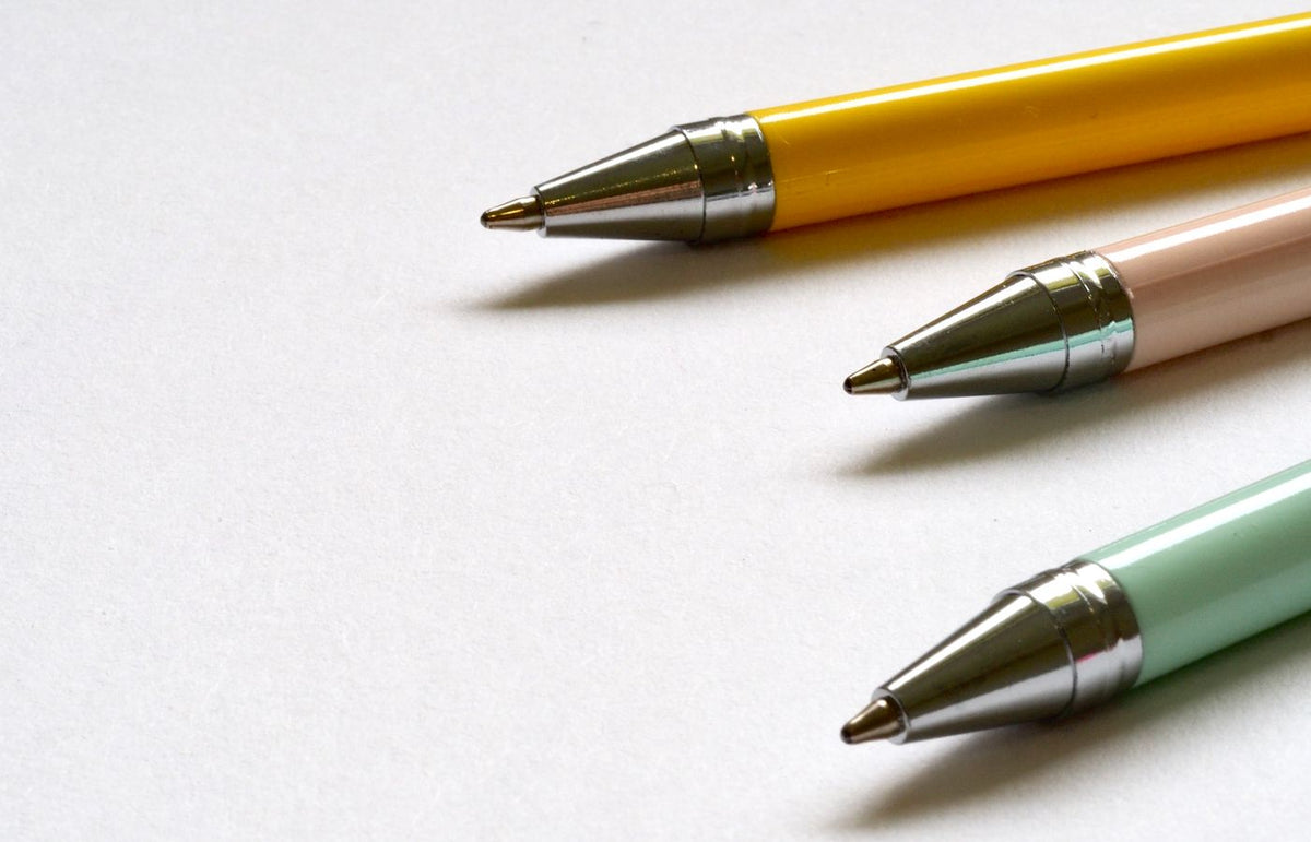 Three multicolored ballpoint pens lay on a clean white surface in a minimal style.