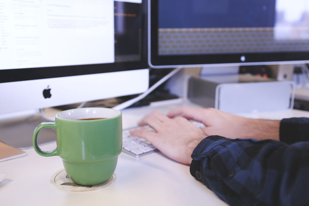 Person typing at a desk with coffee and computer screens. Perfect for business or technology themes.